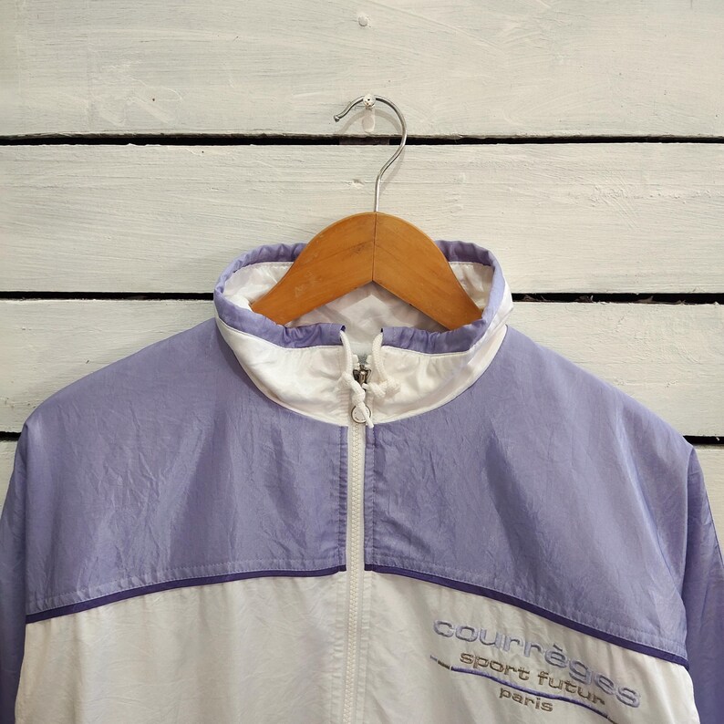 May include: A light purple and white windbreaker jacket featuring a high collar with a front zipper closure. The chest area displays the embroidered text "courr&egrave;ges sport futur paris" in a dark purple thread on the white lower section.