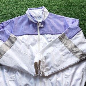 May include: A lightweight windbreaker jacket featuring a color-blocked design in lavender, white, and grey. The jacket has a high collar with a drawstring, a full-length front zipper, and elasticized cuffs. The text "court" is printed on the chest.