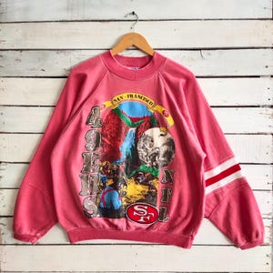Może przedstawiać: W vintage różowej bluzie z nadrukiem wodospadu i napisem "SAN FRANCISCO" w banerze. Bluza ma biało-czerwone paski na rękawach oraz logo "49ers NFL".
