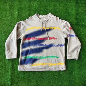 May include: A heather gray long-sleeve sweatshirt with a mock neck and drawstring. The front features three diagonal dark blue spray-painted stripes overlaid with repeating text that reads Formula Racing Benetton Uno in yellow, red, blue, and green.