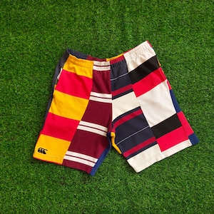 May include: Colorful patchwork shorts with a mix of red, yellow, white, navy, and burgundy panels. The shorts have a small logo on the left leg and a blue trim. The shorts are made of a soft material.
