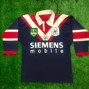 May include: A navy blue long-sleeved jersey with red and white detailing. The jersey displays the text "SIEMENS mobile" in white, alongside team logos and "Sydney City Roosters". The collar is white.