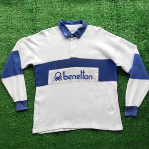 May include: A long-sleeve rugby shirt in white and blue with a blue collar and cuffs. The shirt features a wide horizontal blue stripe across the chest with the text "benetton" printed in blue, accompanied by a blue logo icon to the left.