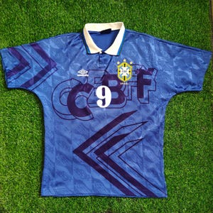 Könnte beinhalten: Blaues Brasilien-Fußballtrikot mit der Nummer 9 auf der Vorderseite. Das Trikot hat einen weißen Kragen und das CBF-Logo. Das Umbro-Logo befindet sich auf der Brust. Das Trikot hat ein geometrisches Muster.