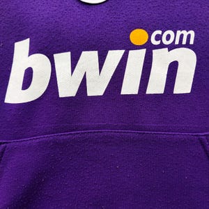 May include: Close-up of a purple hoodie with the white text "bwin" and a yellow dot with the word "com". The hoodie has a pocket at the bottom and a textured surface.