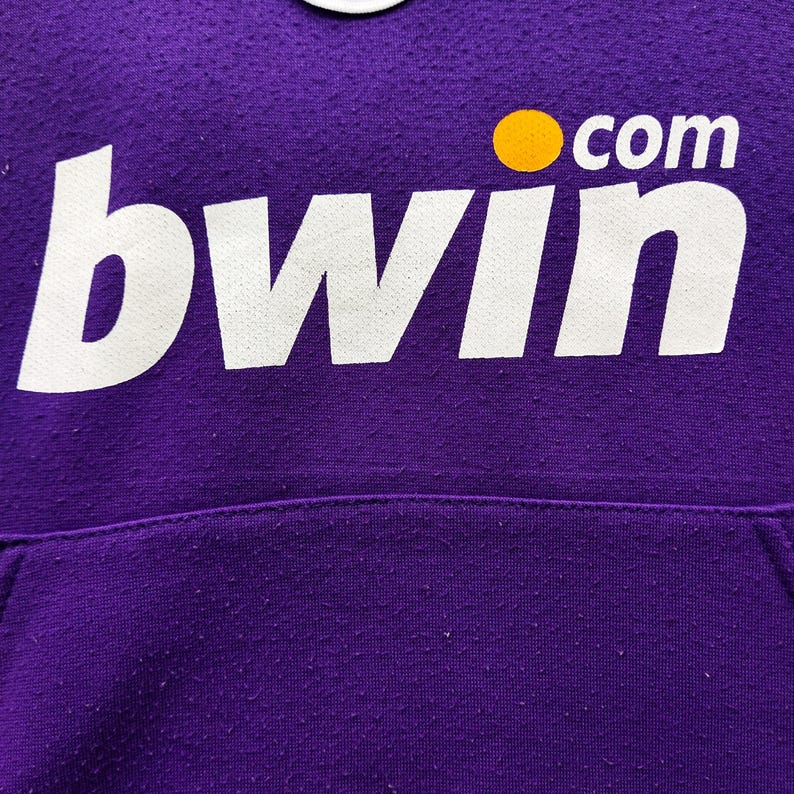 May include: Close-up of a purple hoodie with the white text "bwin" and a yellow dot with the word "com". The hoodie has a pocket at the bottom and a textured surface.