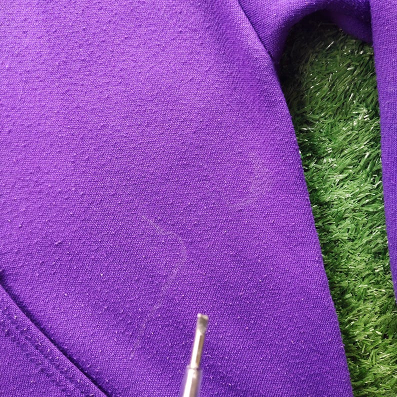 May include: Close-up of a textured, purple fabric, possibly a sweatshirt or hoodie. The fabric has a slightly worn appearance with small, raised fibers. A small, silver tool is visible at the bottom, suggesting a repair or inspection.