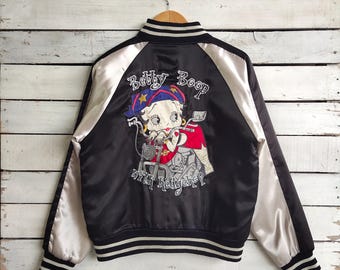 Può includere: Giacca bomber in raso nero con maniche bianche e grafica di Betty Boop sul retro. La grafica presenta Betty Boop in sella a una moto con la scritta "Betty Boop with Hedgehog". La giacca ha colletto e polsini a righe.