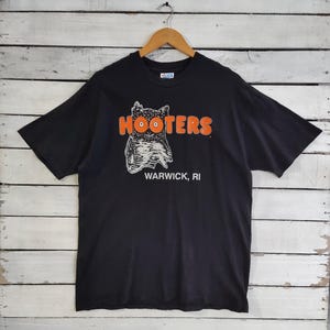 May include: Black t-shirt with the word "HOOTERS" in orange letters above a white and gray owl graphic. Below the graphic, the text "WARWICK, RI" is printed in black. The shirt is hanging on a wooden hanger.