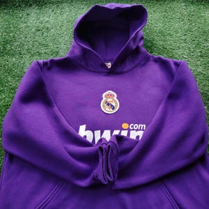 May include: A purple hoodie with a hood and a front pocket. The hoodie features a team logo patch and the text "bwin.com" in white and orange. The hoodie is made of a soft material and is likely for casual wear.