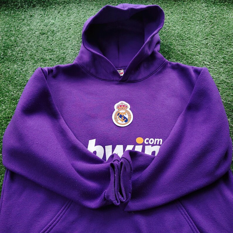 May include: A purple hoodie with a hood and a front pocket. The hoodie features a team logo patch and the text "bwin.com" in white and orange. The hoodie is made of a soft material and is likely for casual wear.