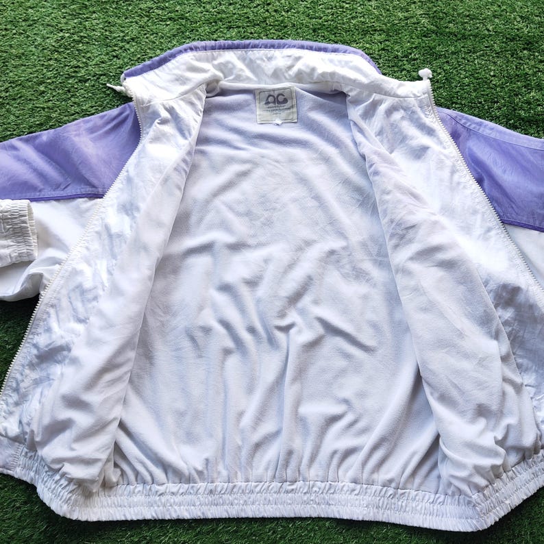May include: A white and light purple windbreaker jacket with a full-length front zipper, long sleeves, and elastic cuffs. The interior is lined with white fabric and features a rectangular brand label at the neck with the text AG.