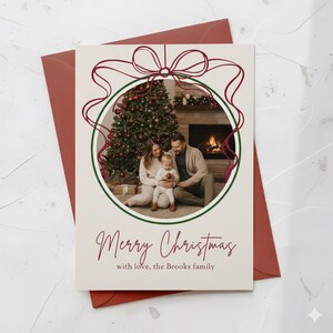 Pode incluir: Cartão de Natal com uma foto de família dentro de um círculo verde, adornado com um design de fita bordô. O cartão diz "Merry Christmas" e o sobrenome. O cartão está em um envelope bordô.