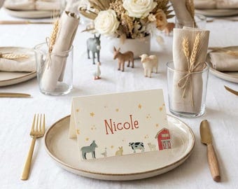 May include: A table setting with a beige plate, gold fork and knife, and a place card that reads "Nicole." The place card has illustrations of farm animals and a red barn. Clear glasses hold rolled napkins and wheat stalks.