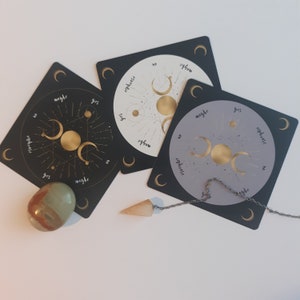 May include: Three black and white circular divination cards with gold moon phases and the words "yes", "no", "maybe", and "rephrase" printed on them. A silver chain with a clear quartz pendulum is hanging over the cards.