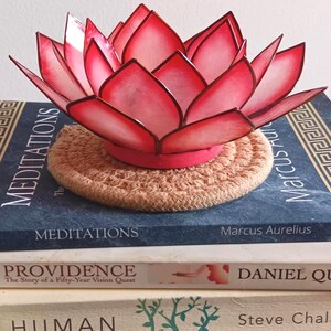 Puede incluir: Una escultura decorativa de flor de loto en tonos rojos y rosas se encuentra sobre una pila de libros. Los libros incluyen títulos como "Meditaciones". La escultura descansa sobre un posavasos circular tejido.