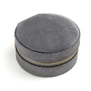 Puede incluir: Un estuche redondo de joyería de terciopelo gris con una cremallera dorada. El estuche está cerrado y sobre un fondo blanco. El material de terciopelo tiene un aspecto suave y texturizado.