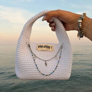 Puede incluir: Un bolso de mano blanco tejido a ganchillo con una sola asa, que presenta una etiqueta de tela beige con el texto "menta" y un icono de rayo. El bolso está decorado con una cadena plateada superpuesta y una tira de cuentas multicolor con dijes.