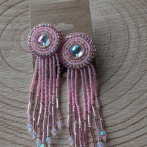 May include: Pink beaded earrings with a circular design and dangling strands. The earrings feature a central iridescent gem surrounded by concentric circles of pink beads and smaller iridescent beads. The dangling strands end with iridescent crystal beads.