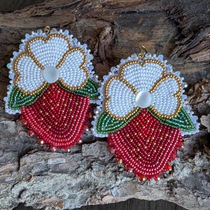 May include: A pair of beaded strawberry earrings. Each earring features a red strawberry base with white beaded flower tops, gold accents, and green leaves. The earrings are set against a natural wood background.