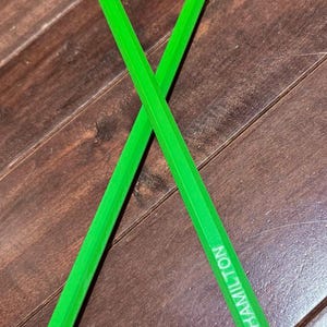 Puede incluir: Dos baquetas verdes brillantes cruzadas sobre una superficie de madera. El texto "RIGGS HAMILTON" está impreso en una de las baquetas. Las baquetas son de un verde vibrante.