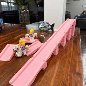 Könnte beinhalten: Rosa Spielzeugbrücken- und Rampenset mit zwei Spielfiguren, die auf weißen Pferden reiten. Das Set besteht aus Kunststoff und steht auf einem Holztisch. Ein Strauß roter Rosen befindet sich im Hintergrund.