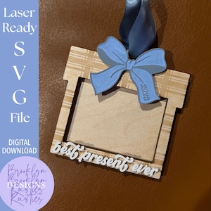 Op de afbeelding: Een houten cadeau-vormig ornament met een lichtblauwe strik en lint. Het ornament heeft een rechthoekig frame met de woorden "best present ever" in witte letters. Het jaar "2023" staat op de strik. De tekst "Laser Ready SVG File DIGITAL DOWNLOAD" staat links.
