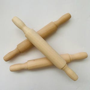 May include: Three wooden rolling pins are arranged on a white surface. The rolling pins are made of light-colored wood and have a classic design with a cylindrical rolling surface and rounded handles. They are ideal for baking.