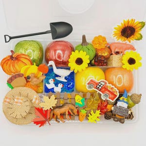 May include: A clear plastic container filled with fall-themed decorative items. Includes pumpkins, sunflowers, a pig, a truck, a shovel, and other autumn-themed embellishments. The container is filled with various colors and textures.