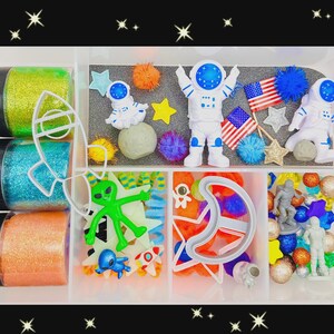 May include: A clear plastic container filled with space-themed craft supplies. Includes glitter, astronaut figures, star and moon-shaped cookie cutters, pom-poms, and small American flags. The glitter is in green, blue, and orange containers.