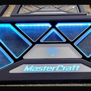 May include: Black panel with blue LED lighting, featuring geometric shapes and the text "MasterCraft" in a light blue font. The panel has a central emblem and is likely a component of a vehicle's interior.