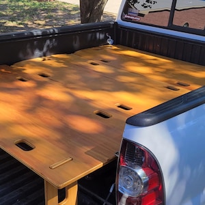 May include: A light brown wooden platform bed frame with rectangular cutouts installed in the cargo bed of a white pickup truck. The flat, modular wooden surface spans the width of the truck bed, supported by vertical wooden legs.
