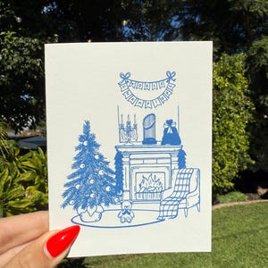 May include: A blue and white Christmas-themed greeting card. The illustration depicts a decorated Christmas tree, a fireplace with a banner, a chair, and a teddy bear. The card is held by a person with red nail polish.