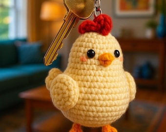 May include: A handmade, yellow crochet chick keychain with a red comb and orange feet. The chick is attached to a silver keyring with a gold key. The chick has black eyes and a small orange beak. The keychain is a cute accessory.