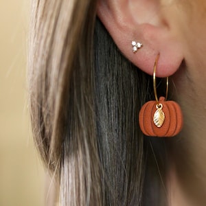 Puede incluir: Un par de pendientes de aro dorados con un pequeño dije de calabaza. La calabaza es naranja con un detalle de hoja dorada.