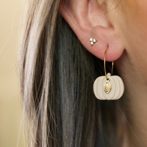 Puede incluir: Un par de pendientes de aro dorados con un pequeño encanto de calabaza blanca que cuelga de uno de los pendientes. El encanto de calabaza tiene un pequeño detalle de hoja dorada. Los pendientes se llevan en la oreja de una persona.