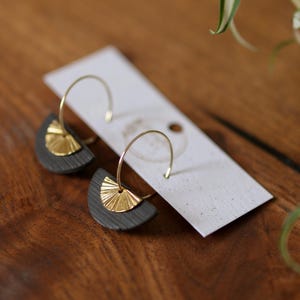 May include: A pair of gold hoop earrings with a dark gray, semi-circular base and a gold sunburst design. The earrings are displayed on a white card, set against a wooden surface with a plant in the background.