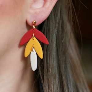 May include: A pair of dangle earrings with three teardrop-shaped charms. The top charm is red, the middle charm is yellow, and the bottom charm is white. The earrings are made of polymer clay and have gold-colored hooks.