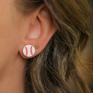 May include: A round stud earring featuring a white baseball design with red stitching, accompanied by a small gold-colored star-shaped stud earring. The baseball earring is circular with a smooth finish and the star earring is a simple metallic shape.