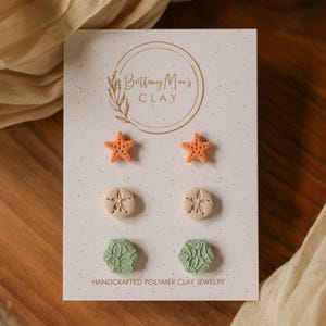 May include: Three pairs of stud earrings on a white display card labelled Brittany Mae's CLAY. The top pair are orange starfish, the middle pair are beige sand dollars, and the bottom pair are sage green hexagons with a floral texture pattern.