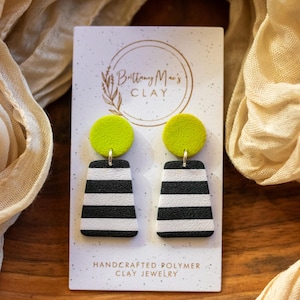 May include: A pair of handmade polymer clay earrings with black and white striped geometric shapes and bright green round accents. The earrings are displayed on a white card with the text "Brittany Mae's CLAY" and "HANDCRAFTED POLYMER CLAY JEWELRY".