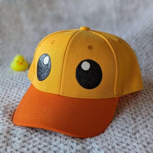 Pode incluir: Boné de beisebol amarelo com aba laranja, com um design de rosto de pato de desenho animado. O boné tem olhos grandes pretos e brancos, e um pequeno pato de borracha amarelo no fundo. Um acessório divertido.