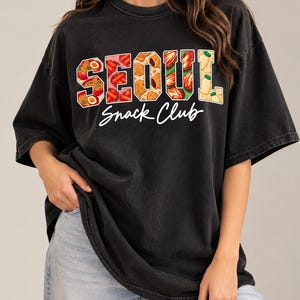 Puede incluir: Una camiseta negra de manga corta con el texto SEOUL en letras grandes de bloque rellenas con ilustraciones de varios aperitivos y alimentos coreanos, con las palabras Snack Club escritas en cursiva blanca debajo del texto principal.