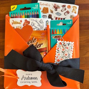 May include: An orange envelope tied with a black ribbon and a tag labeled Cozy Autumn coloring book. Inside are 12 coloring pencils, a pack of 8 Bic crayons, a sheet of autumn-themed stickers, a coloring page, and a packet of apple cider mix.