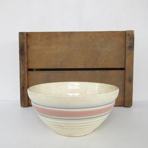 May include: A white ceramic mixing bowl with blue and pink stripes. The bowl has a ribbed design and is sitting on a wooden crate.