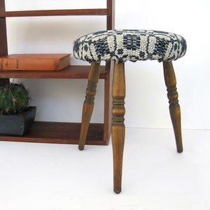 May include: A round stool with a padded seat featuring a black and white geometric woven pattern. The stool has three turned wooden legs with a dark brown finish. The seat is circular and upholstered in a textured fabric with a repeating design.