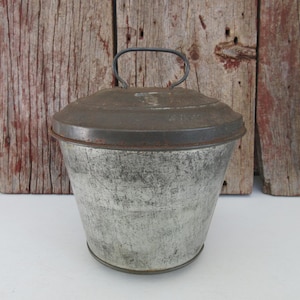 May include: A vintage metal bucket with a lid and handle. The bucket has a silver finish with a distressed appearance, and the lid and handle are a darker, aged metal. The background is a rustic wooden wall.