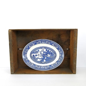 May include: An oval ceramic platter featuring a blue and white willow pattern. The design depicts a landscape with a bridge, trees, and a pagoda, surrounded by a decorative border of geometric shapes and floral motifs on a white background.