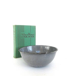 May include: A gray enamelware bowl with a speckled pattern, next to a vintage green cookbook titled "The American Woman's Cook Book." The bowl has a dark rim and a classic design, perfect for kitchen decor.