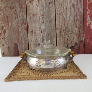 May include: A vintage silver casserole dish with a clear glass lid. The dish has decorative cutouts and orange-toned handles. It sits on a woven, triangular placemat. The background is a weathered wooden wall.